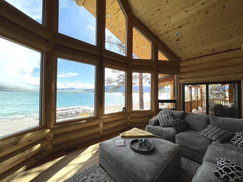 A cozy log home living room in a log cabin featuring large windows, a television mounted on the wall, and a dog resting on a couch. A telescope is positioned near the window with views of trees and an outdoor landscape.