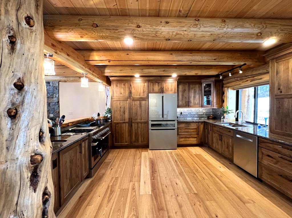 A rustic log home kitchen with wooden cabinets, a large wooden pillar, stainless steel appliances, and a spacious hardwood floor.