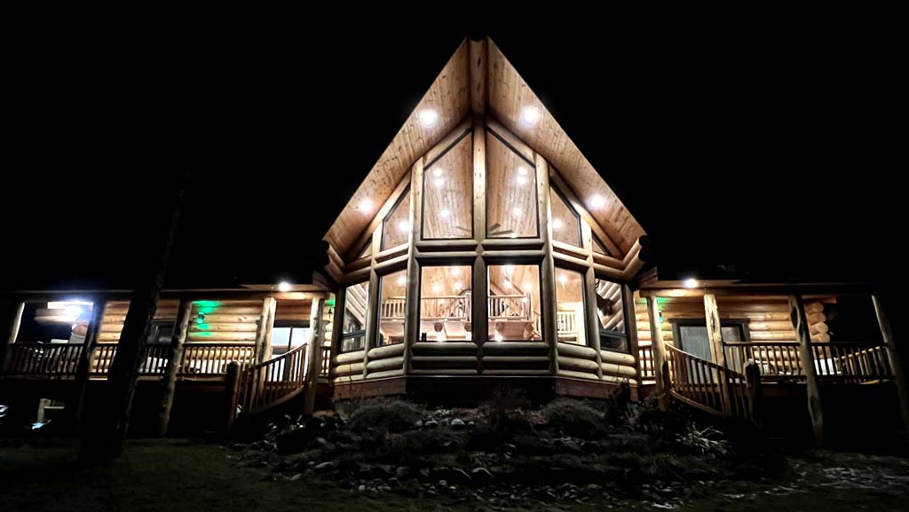 A log cabin illuminated at night, showcasing a large A-frame design with multiple windows and a wooden deck.