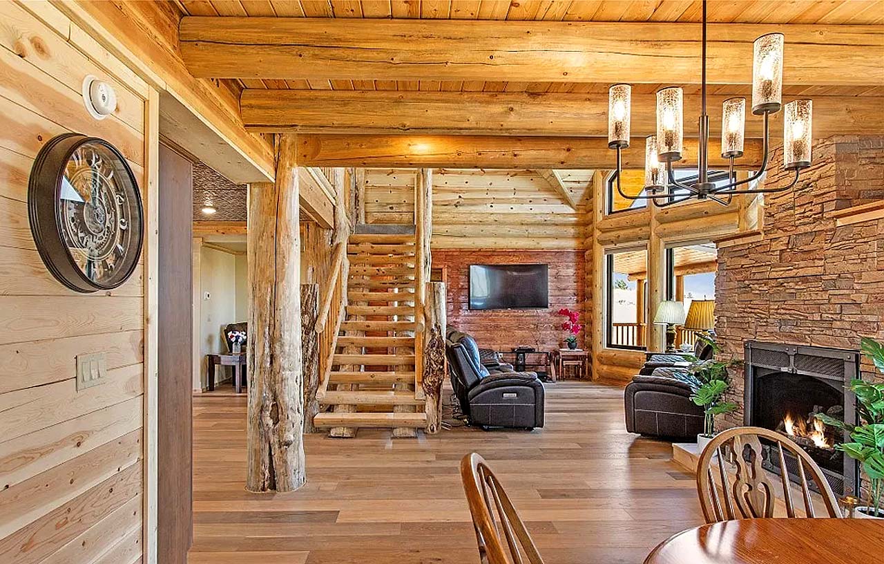 Interior of a rustic log home with wooden beams, a staircase, stone fireplace, and dining area featuring wooden furniture. Large windows allow natural light to flood the space.