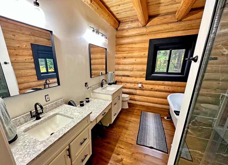 A modern log home bathroom with two sinks, large mirrors, a walk-in shower, and a freestanding bathtub. The walls are adorned with wooden logs, creating a rustic feel.