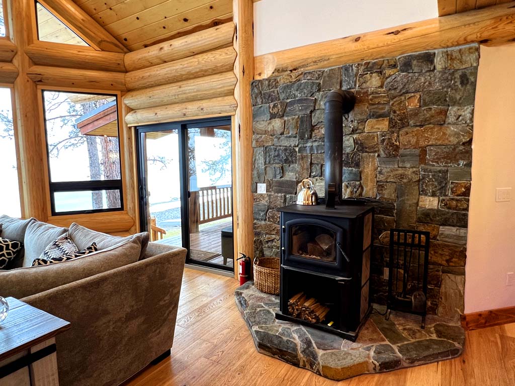 A cozy log home living room with log cabin architecture featuring a stone fireplace, wooden ceiling, and large windows overlooking the outdoors.