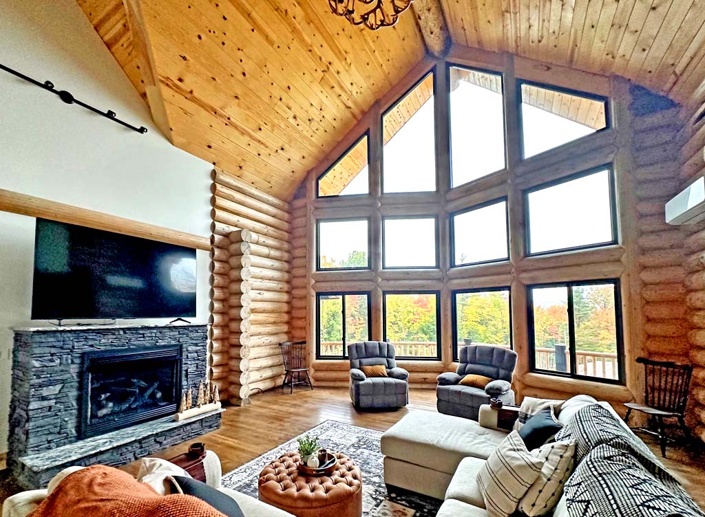 A spacious log home living room with high wooden ceilings, large windows showcasing fall foliage outside, a stone fireplace, a sectional sofa with decorative pillows, and comfortable armchairs.