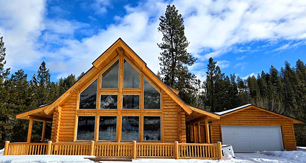A large wooden cabin with a steeply pitched roof, featuring multiple large windows and a front porch. Snow covers the ground around the cabin, and tall pine trees are visible in the background under a partly cloudy sky.