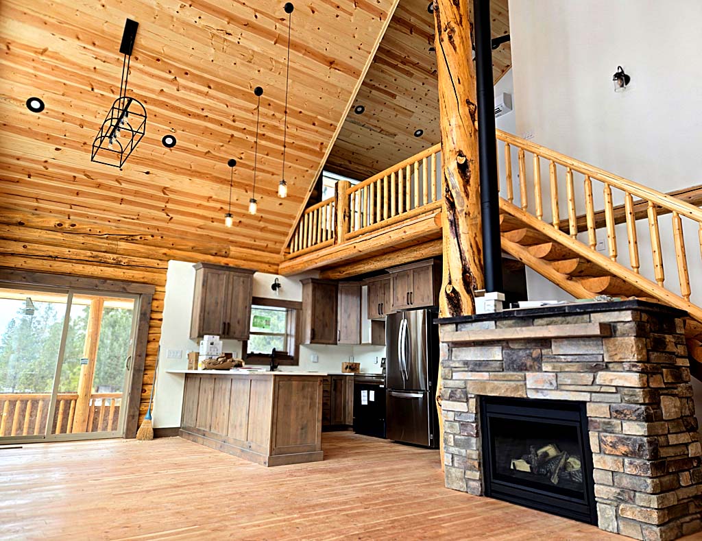 Interior view of a modern log home, featuring wooden beams, a stone fireplace, and an open staircase with wooden railings. Large windows let in natural light, and the kitchen has wooden cabinets and stainless steel appliances.