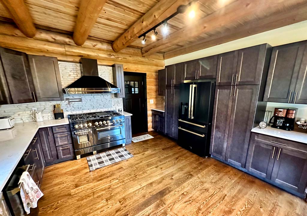 A modern log home kitchen featuring dark wooden cabinetry, a stainless steel stove with a range hood, and a marble countertop. The floor is made of hardwood, and there is a door leading to another room. The kitchen has various appliances including a refrigerator.