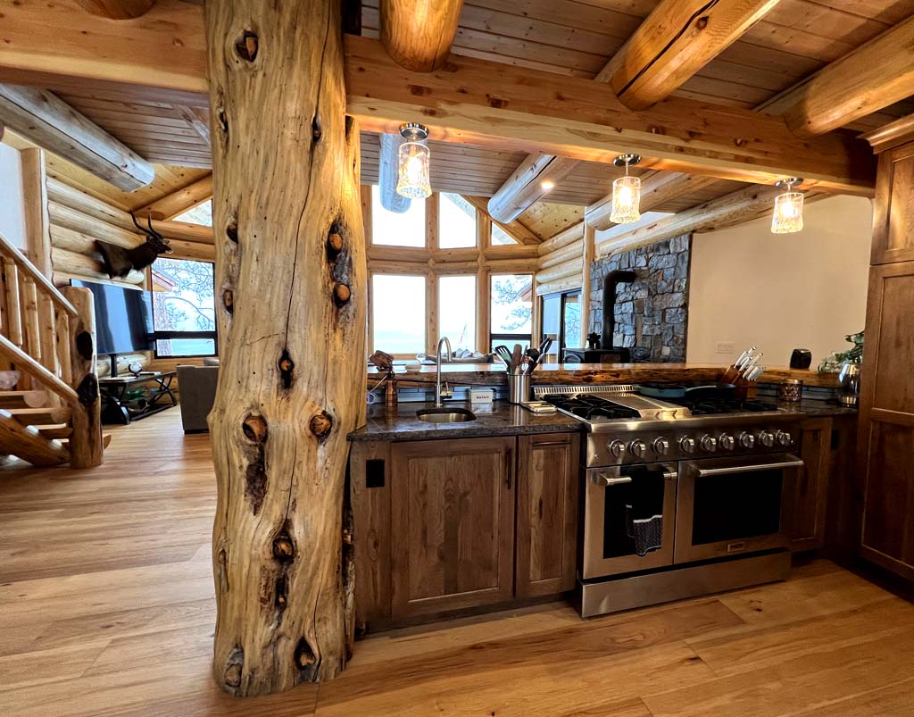 A rustic log home kitchen with wooden cabinetry, a large stove, and a view of an open living space featuring log beams and large windows.