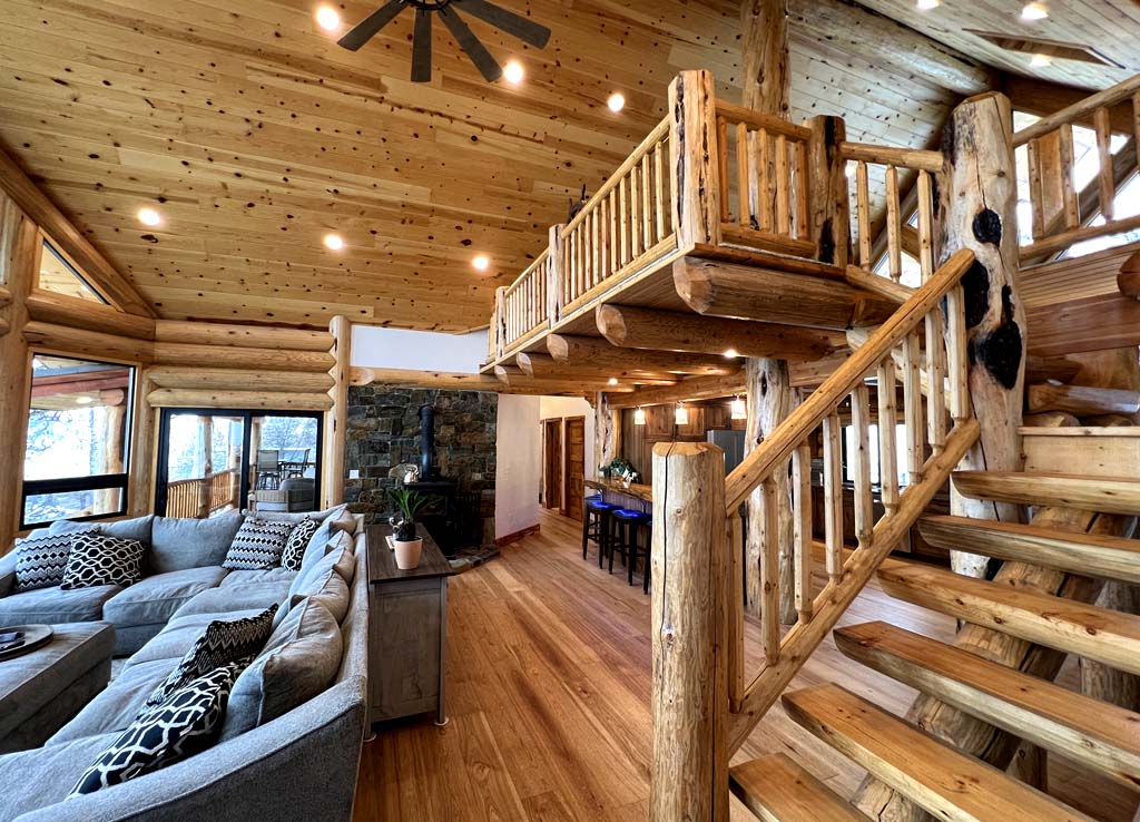 A cozy log home interior featuring a wooden staircase, a large sectional sofa, and plenty of natural light. The walls and ceiling are made of polished wood, creating a warm atmosphere.