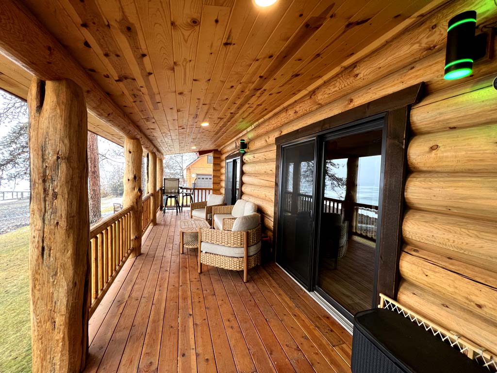 A wooden porch with log walls, featuring two wicker chairs and a sliding glass door, surrounded by a scenic view.