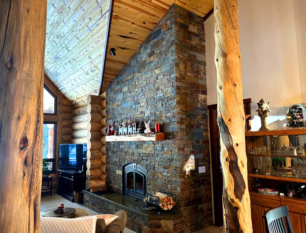 Interior of a cozy log home featuring a stone fireplace, wooden beams, and large windows.