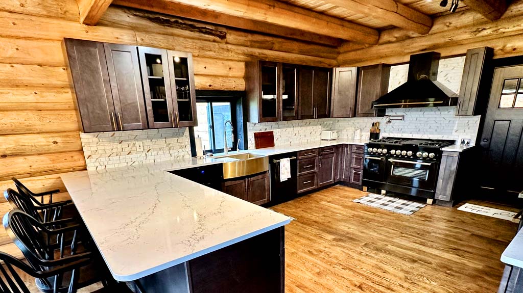 A modern log home kitchen with dark wood cabinets, a large island with white countertops, stainless steel appliances, and a rustic log cabin-style interior Home kitchen design Lazarus BED 1024