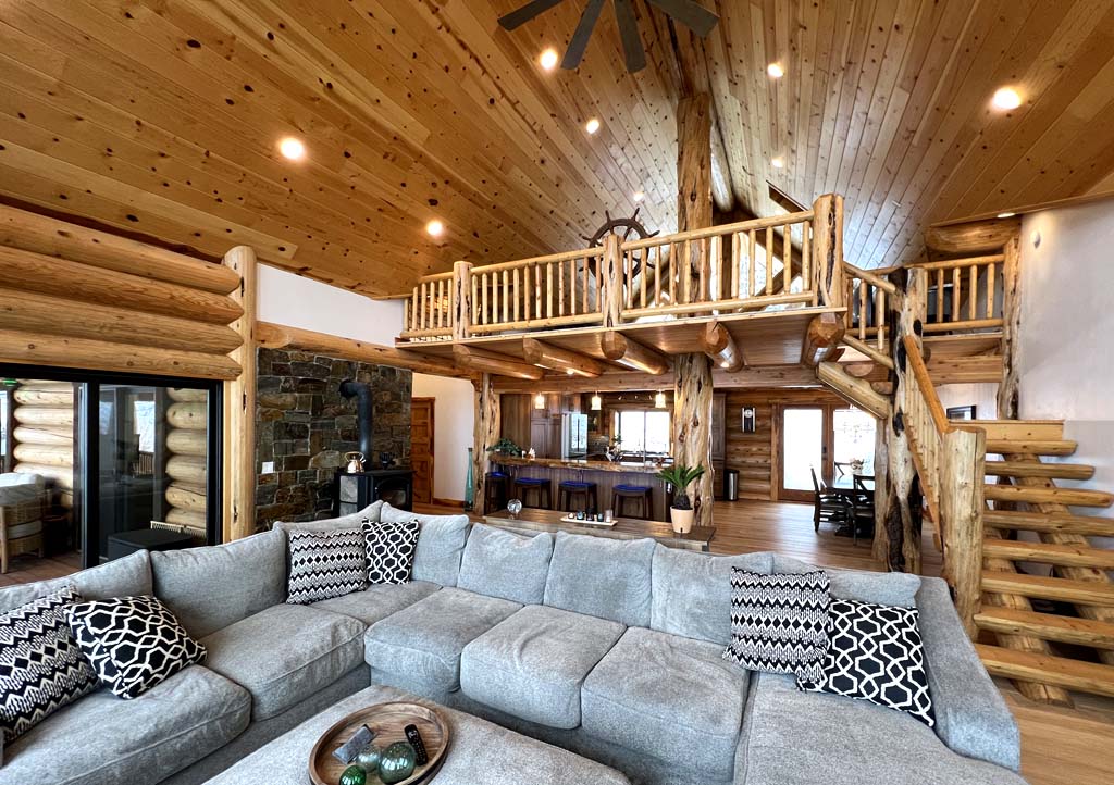 A spacious log home interior featuring a large gray sectional sofa with decorative pillows, wooden ceiling beams, and a staircase leading to an upper level. The space is well-lit with natural light and includes a stone wall and wooden accents throughout.