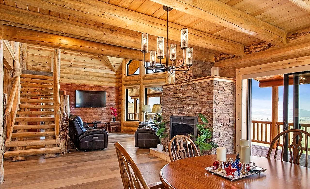 A cozy, wooden log home interior of a log cabin featuring a dining area with a round table, chairs, and candles. In the background, there is a staircase leading to an upper level, large windows allowing natural light in, and comfortable seating arrangements. A stone fireplace adds to the rustic charm of the room.