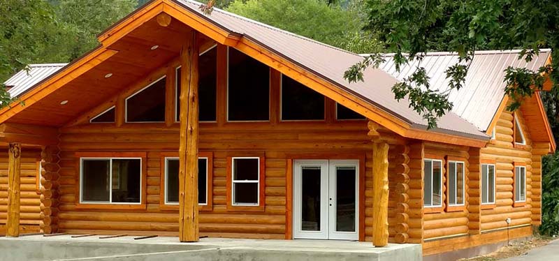 A large log cabin with a sloped roof, featuring large windows and a porch area.