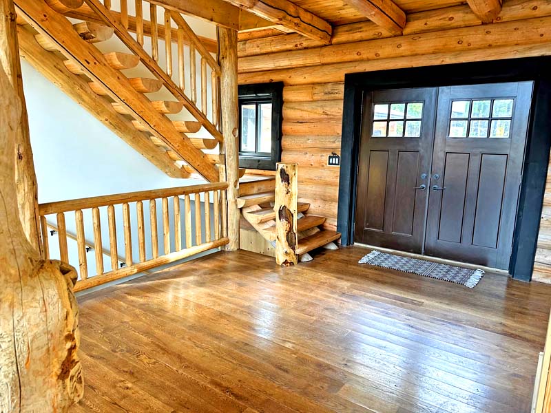 A wooden log home interior featuring a staircase, wooden railings, and double front doors. The floor is made of polished wood, and there is a small welcome mat by the entrance.