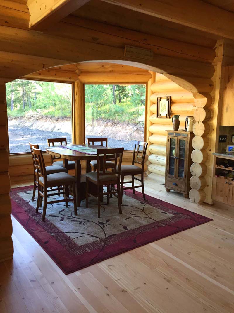 A cozy wooden dining area with a round table surrounded by chairs, set on a rug. Large windows allow natural light and a view of greenery outside. The walls are made of log wood, and there’s a small cabinet in the corner.