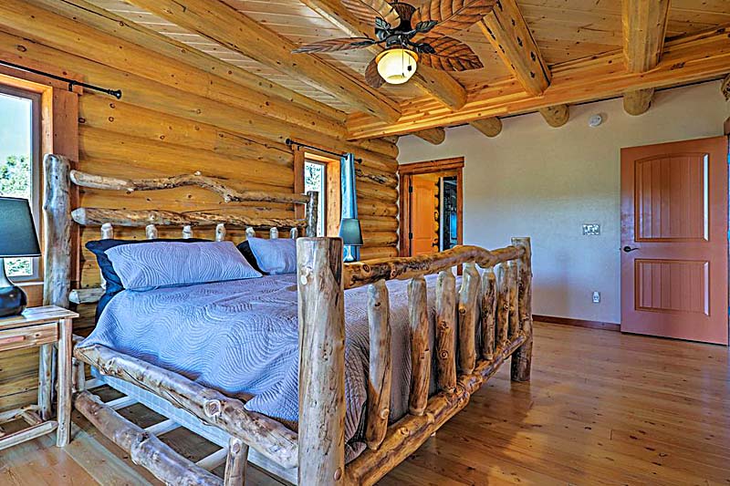 A rustic log home bedroom featuring a wooden bed frame, a blue and gray bedding set, and exposed wooden beams on the ceiling. There is a nightstand with a lamp beside the bed and a wooden door leading to another room.