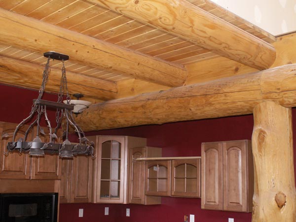 A wooden beam ceiling in a room with a chandelier, featuring log-style pillars and light-colored cabinetry against a dark red wall.