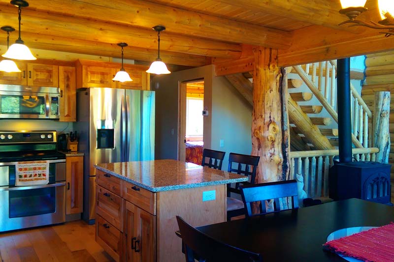A cozy log home kitchen area in a log cabin, featuring wooden cabinets, stainless steel appliances, a large island with a granite countertop, and a staircase made of logs. The dining table is set nearby with black chairs.
