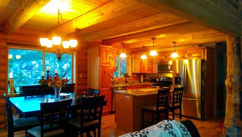 A cozy log home kitchen and dining area in a log cabin, featuring wooden beams, a large table with chairs, cabinets, and modern appliances. Warm lighting enhances the inviting atmosphere.