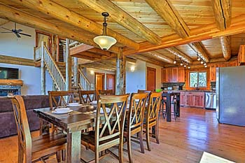 A rustic log home dining area with a wooden table and chairs, featuring exposed beams and a spacious kitchen in the background.