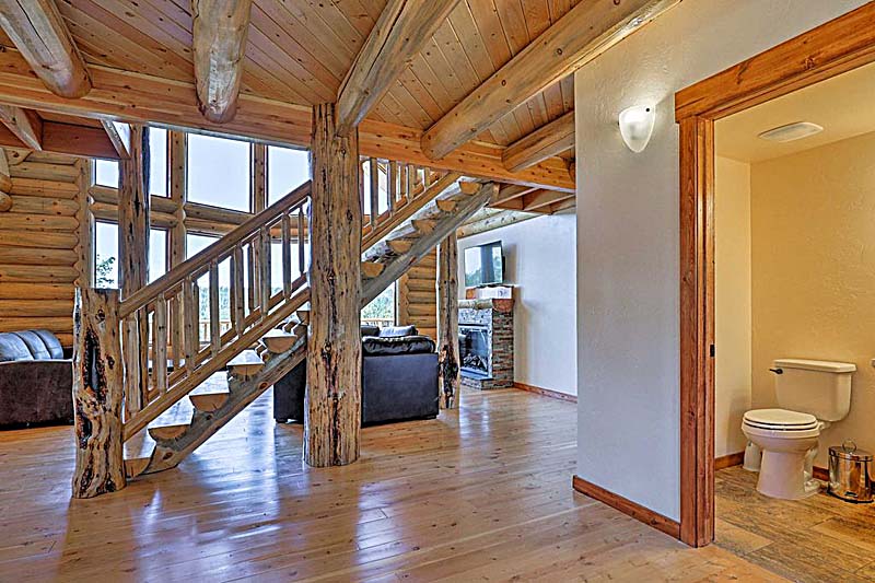 A spacious log home wooden interior with a staircase, large windows letting in natural light, a leather couch, and an open view towards another room featuring a fireplace and bathroom.