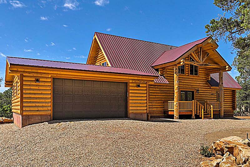 A log home with a steep roof, garage, and porch, set in a gravel area surrounded by trees.