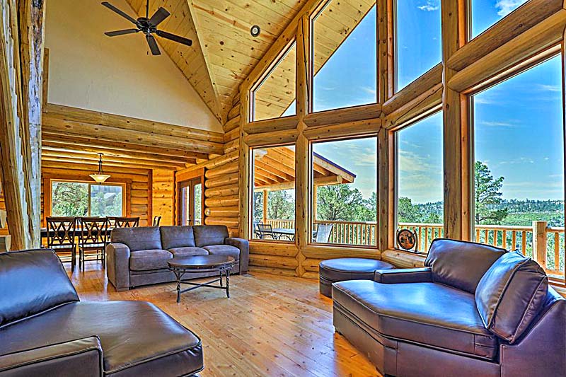 A spacious, modern log home interior with large windows providing a view of the outdoors. The living area features dark leather furniture and wooden accents, creating a warm and inviting atmosphere.