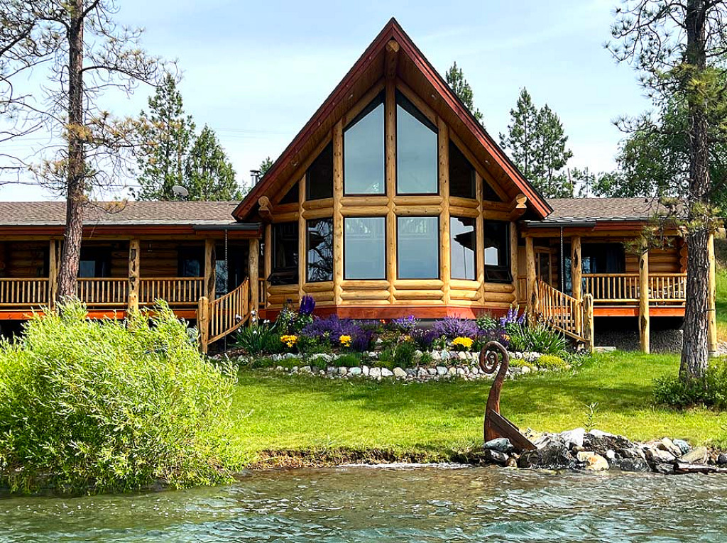 A large log cabin with a triangular roof, featuring many windows, surrounded by greenery and colorful flowers near a body of water.