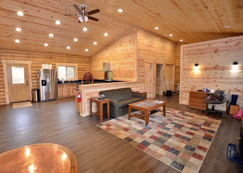 A log home spacious, wooden interior of a cabin with a kitchen and living area, featuring a couch, coffee table, and a multi-colored rug on the floor. There are light fixtures in the ceiling and large windows allowing natural light to enter.