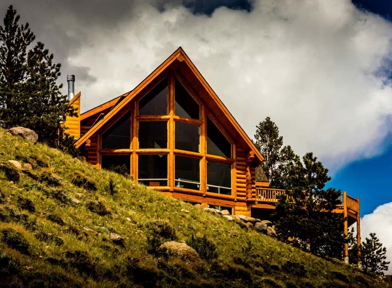 A beautiful wooden cabin with large windows, situated on a grassy hillside with trees in the background and a cloudy sky above.