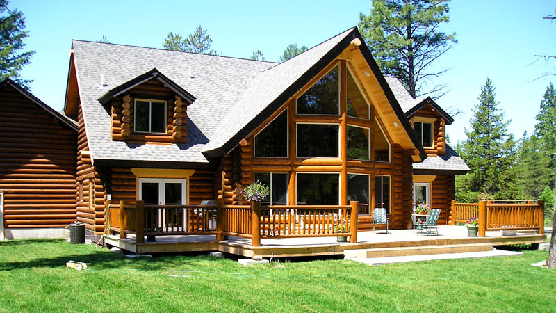 A large log cabin with a wooden deck surrounded by trees and grass. The cabin features large windows, a pitched roof, and an inviting outdoor space.