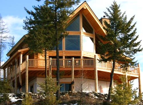 A large wooden house with a modern design, featuring a sloped roof, expansive windows, and a wraparound balcony. The surrounding area includes trees and rocky terrain.
