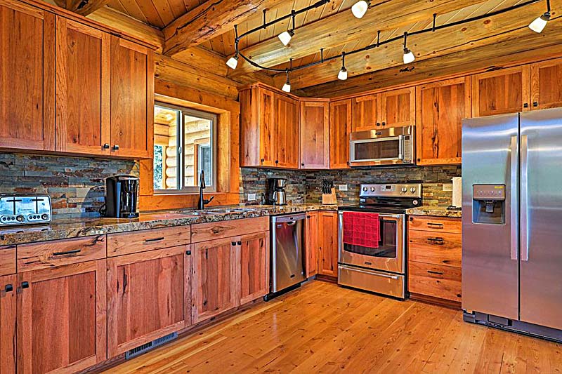 A modern log home kitchen featuring wooden cabinets, stainless steel appliances, and a granite countertop. The space has a warm, rustic feel with exposed wooden beams on the ceiling and soft lighting from spotlights.