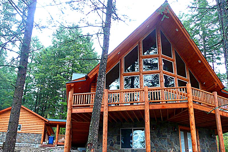 A large log home with a steeply pitched roof, featuring tall windows and a wraparound balcony, surrounded by trees.