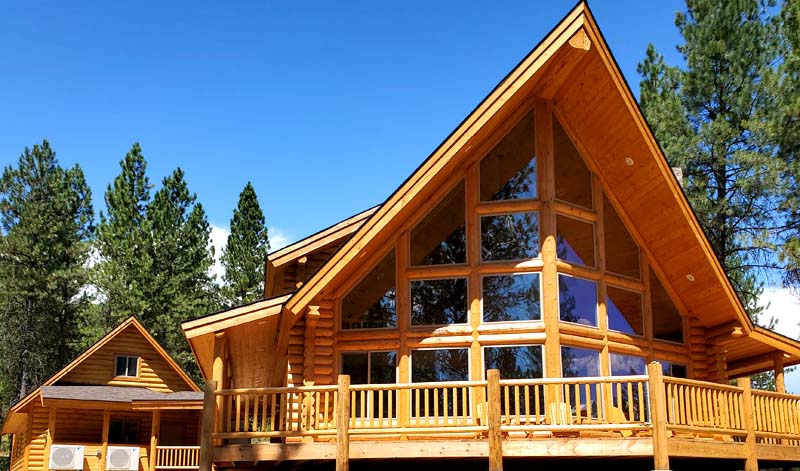 A large wooden cabin with a triangular roof, featuring large windows and surrounded by pine trees under a blue sky.