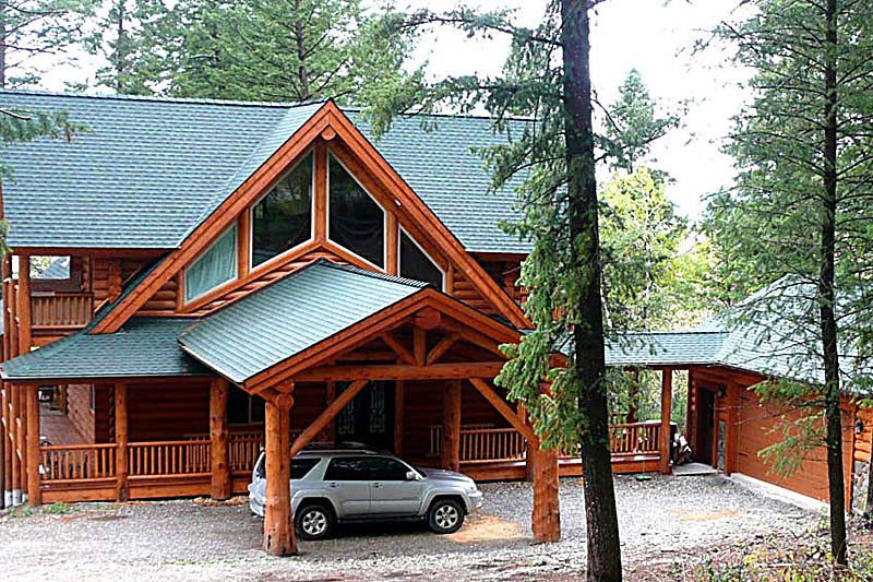 A large log house surrounded by trees, with a vehicle parked in front.