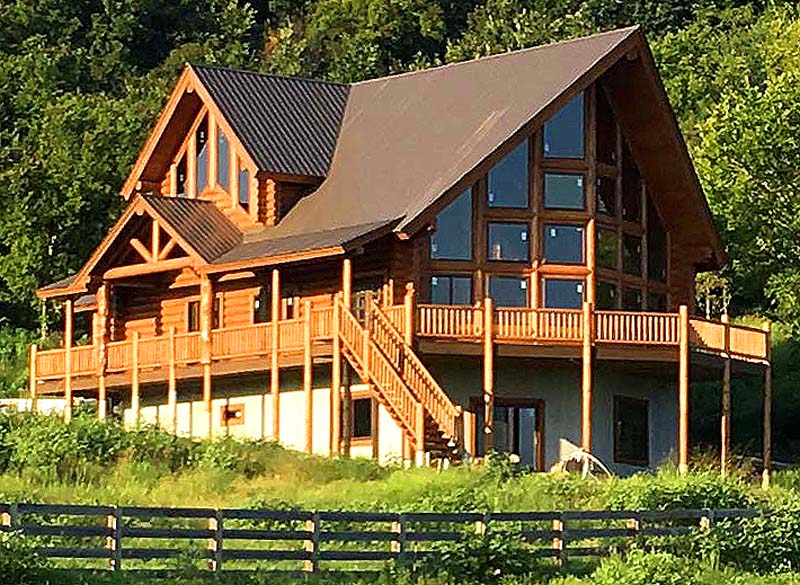 A large, modern log cabin with expansive glass windows and a sloped roof, surrounded by greenery and set against a hillside. The cabin features wooden decks and a spacious entryway.