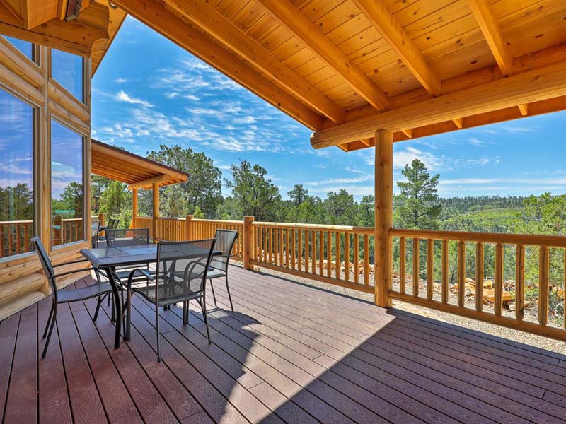 A spacious wooden deck with a dining table and chairs, surrounded by a forested landscape under a clear blue sky.