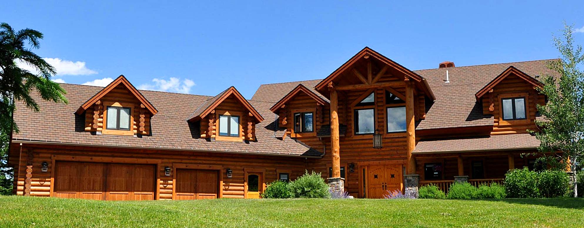 A large log cabin-style house with a brown wooden exterior, multiple gabled roofs, and a spacious front lawn. The house features a central entrance with tall windows and two garage doors on one side.