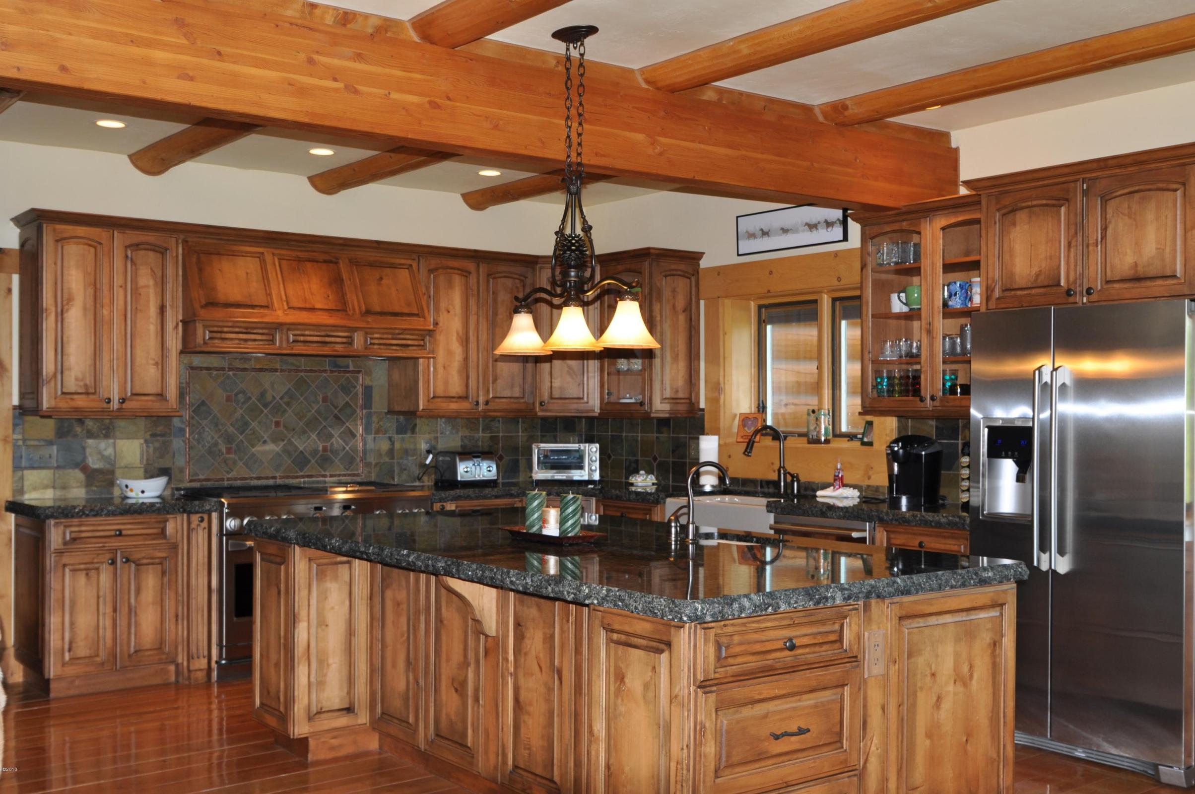 A spacious log home kitchen featuring wooden cabinetry, a large island with a dark granite countertop, stainless steel appliances, and rustic wooden beams on the ceiling.