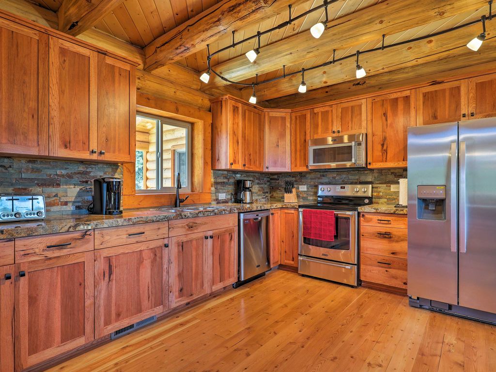 A modern log home kitchen featuring wooden cabinets, stainless steel appliances, and a rustic stone backsplash, with a large window providing natural light.
