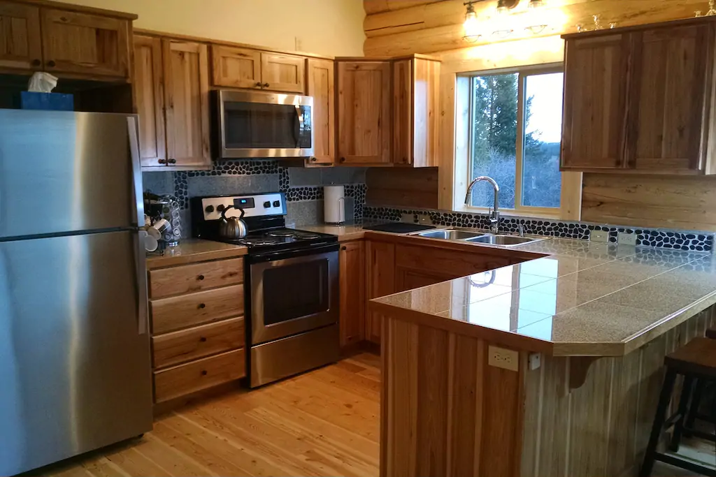 A log home kitchen featuring wooden cabinetry, stainless steel appliances including a refrigerator and stove, and a window letting in natural light. The countertops are tiled, and there is a sink with a modern faucet.