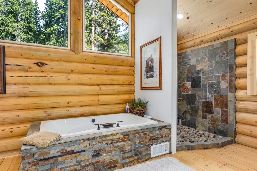 A log home bathroom featuring a bathtub and a shower area with stone walls, set in a log cabin style. Trees are visible through large windows.
