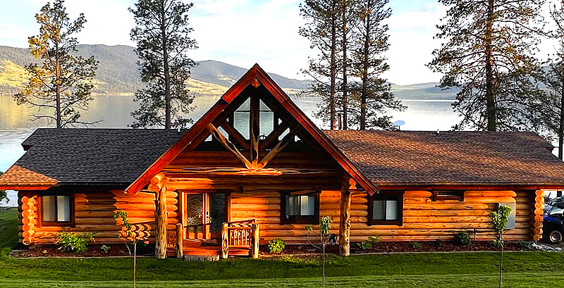 A rustic log cabin surrounded by tall trees, with a lake and mountains in the background. The cabin features a gabled roof and large windows.