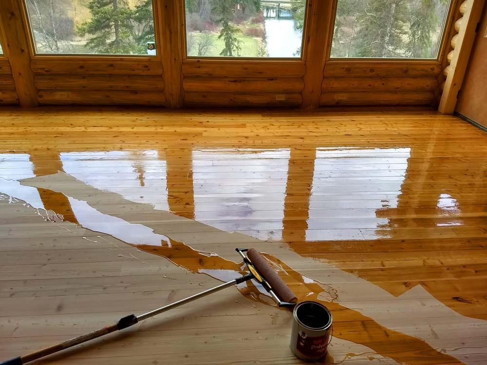 A wooden floor is being freshly coated with a clear finish. A paint roller and can of finish are on the floor, and sunlight is shining through large windows, illuminating the space.