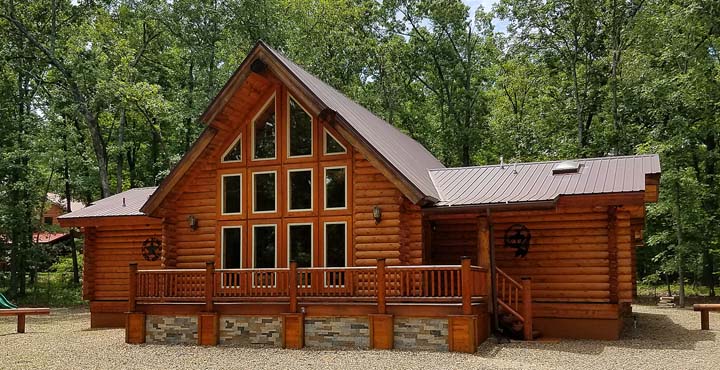A large, rustic log cabin with multiple windows and a peaked roof, surrounded by trees. The cabin features a wooden porch with railings and is set in a gravel area.