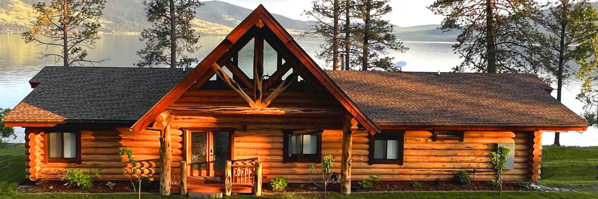 A log cabin with a triangular roof and large windows, surrounded by trees and a body of water in the background.