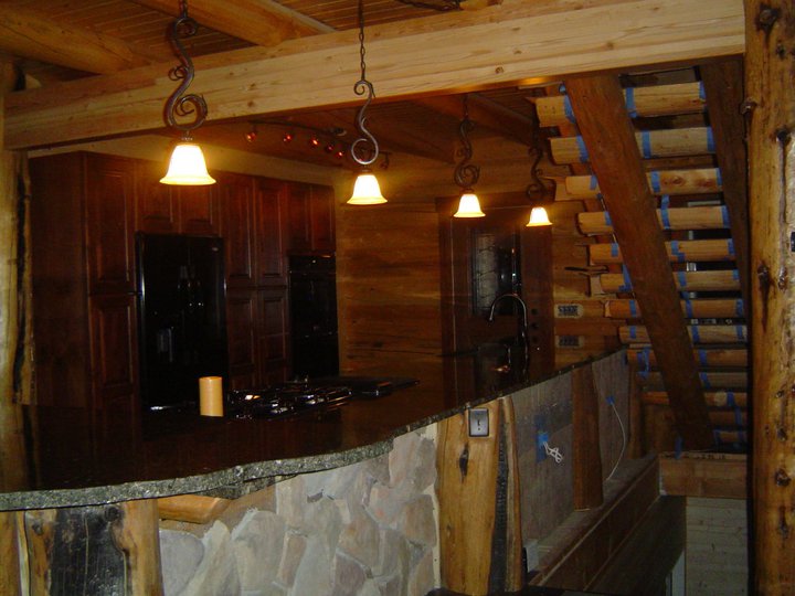 A rustic log home kitchen with wooden beams and cabinetry, featuring a stone bar area and warm lighting.
