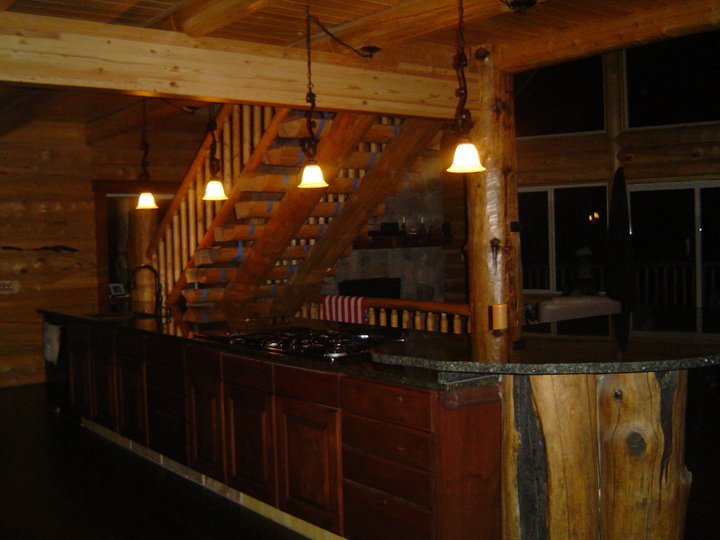 A log home kitchen featuring a wooden bar area, a stove, and pendant lighting. A staircase is visible in the background beside large windows.